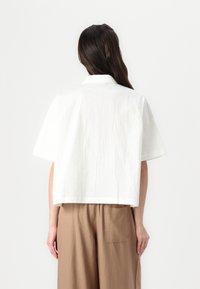 Camisa corta de manga corta blanca con una textura en la tela y un cuello clásico, combinada con pantalones de pierna ancha beige que tienen un bolsillo trasero.