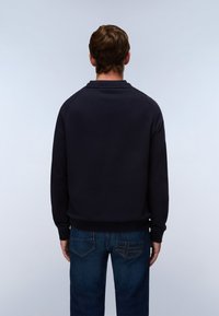 Homme portant une chemise noire à manches longues et un jean bleu, debout dos à un fond gris clair uni.