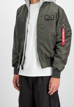 Person iført en åben olivengrøn Alpha Industries bomberjakke over en grå hættetrøje og hvid T-shirt med sorte bukser.