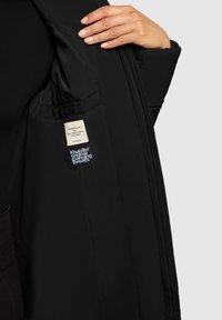 Chaqueta de plumas negra con textura acolchada, que presenta una etiqueta interior y una etiqueta de logo. Cierre de cremallera y ligero aislamiento para mantener el calor.