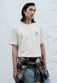 T-shirt di cotone color crema con maniche corte e piccolo motivo grafico nero. Jeans blu con camicia a quadri nera e grigia annodata in vita.