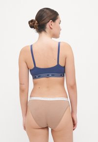 Blå idretts-bh med tynne stropper som har en logo-stripe; paret med beige hipster-truser med en hvit elastisk strikk, vist fra baksiden.