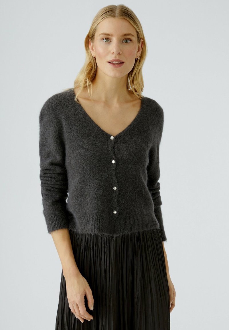 Cardigan en tricot gris foncé avec un col en V, des manches longues et une fermeture à cinq boutons. Texture douce avec un léger éclat des boutons, associé à une jupe plissée.