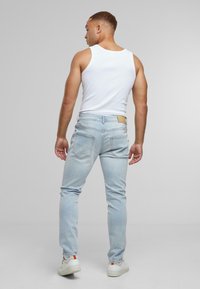 Vyras, vilkintis baltą marškinėlį be rankovių, šviesiai mėlynas aptemptas džinsus ir baltus sportbačius, stovi nugara į kamerą pilkame fone.