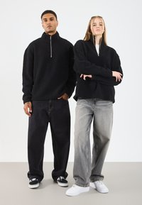 Melnā zip-up polāra džemperis ar struktūru, apvienots ar tumšām brīvām džinsām un baltām sporta apaviem, kā arī ar gaišajiem pelēkajiem džinsiem un baltām kurpēm.