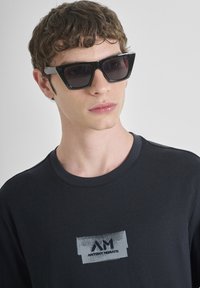 T-shirt en coton noir avec col rond, présentant un écusson gris texturé. Le mannequin porte des lunettes de soleil noires angulaires avec des verres foncés.