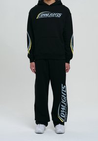 Czarna bluza z kapturem z logo, żółte i niebieskie akcenty na rękawach; w zestawie z czarnymi spodniami dresowymi z nadrukiem logo. Białe buty.