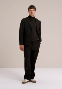 Mann steht mit einem schwarzen Blazer, schwarzem Rollkragenpullover, schwarzen Hosen und weißen Sneakern mit braunen Sohlen vor einem schlichten Hintergrund.