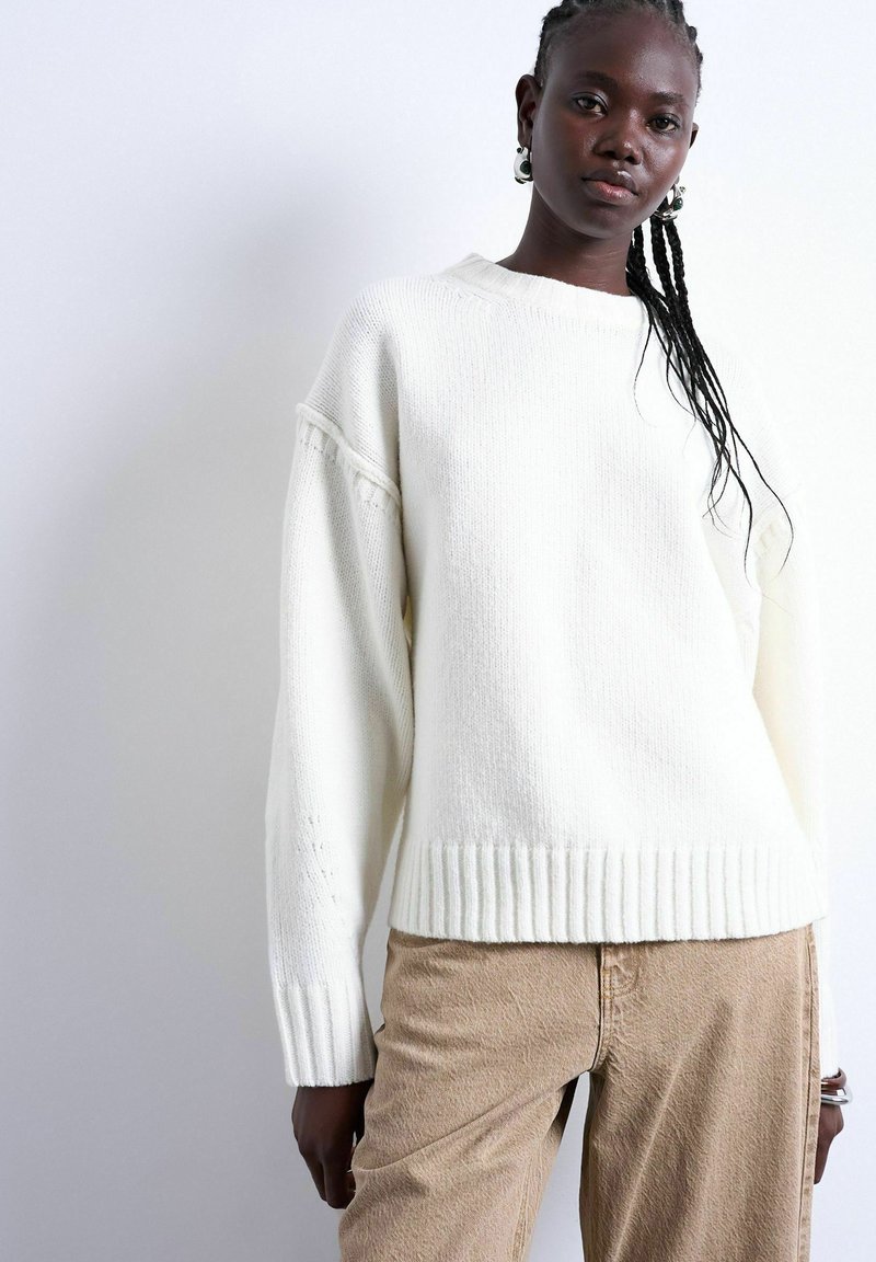 Jeune femme noire aux cheveux tressés portant de grandes boucles d'oreilles anneau, un pull en maille blanc et un pantalon beige sur un fond clair uni.