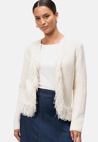 Cardigan blanc texturé avec bord frangé, design ouvert à l'avant, manches longues, porté sur un haut uni, associé à une jupe en denim.