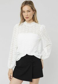Blouse en dentelle blanche avec de longues manches bouffantes et un col haut, présentant un design transparent. Associée à une jupe noire avec des accents de boutons.