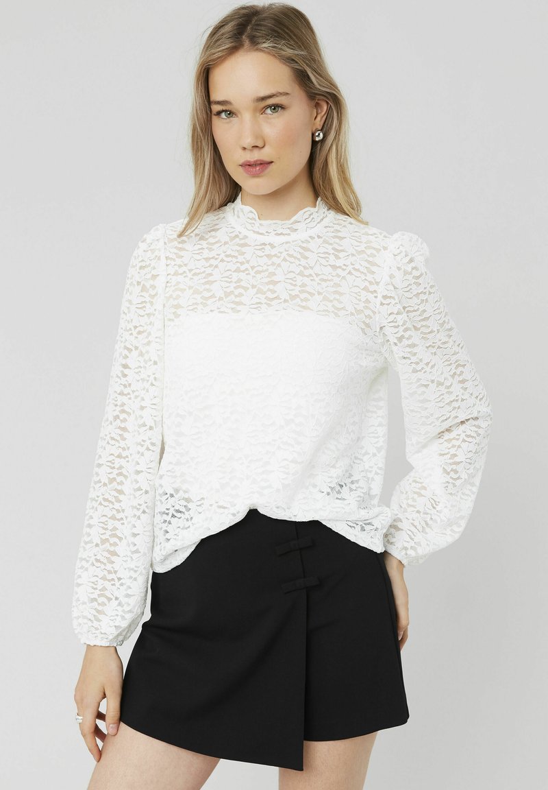 Blouse en dentelle blanche avec de longues manches bouffantes et un col haut, présentant un design transparent. Associée à une jupe noire avec des accents de boutons.