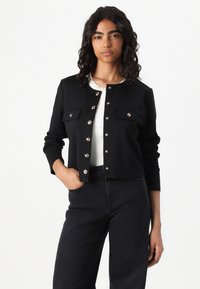 Schwarze, verkürzte Jacke mit goldenen Knopfakzenten, zwei Fronttaschen und rundem Kragen, kombiniert mit einem weißen, gerippten Oberteil und dunklen Jeans.