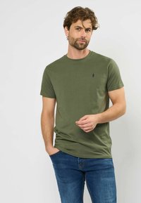 Tamsiai žalia trumpomis rankovėmis megzta marškinėliai su apvalia iškirpte ir maža tamsiai mėlyna emblema krūtinėje, derinami su mėlynos džinsinės kelnės.