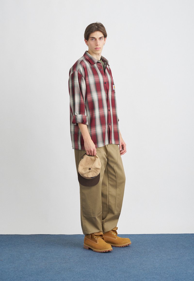 Red and gray plaid shirt, beige pants, tan boots, and a brown-accented cap. Focus on casual outdoor style with varied textures.