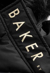 Tela negra con grandes letras beige que forman "BAKER" y un pequeño detalle de piel en la esquina superior derecha.