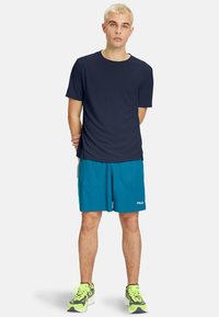 Marineblå kortærmet træningsskjorte parret med teal shorts. Har et FILA-logo på shortsene. Modellen har grå og neon grønne sneakers på.