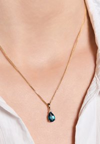 Złoty naszyjnik łańcuchowy z zawieszką w kształcie gruszki z niebieskim kamieniem szlachetnym i srebrnym akcentem, prezentowany na lekko oświetlonym odcieniu skóry.