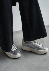 Baskets argentées pailletées avec une semelle blanche �épaisse et un bout rond, équipées de lacets crème et d'œillets en métal, associées à un pantalon noir à jambes larges.