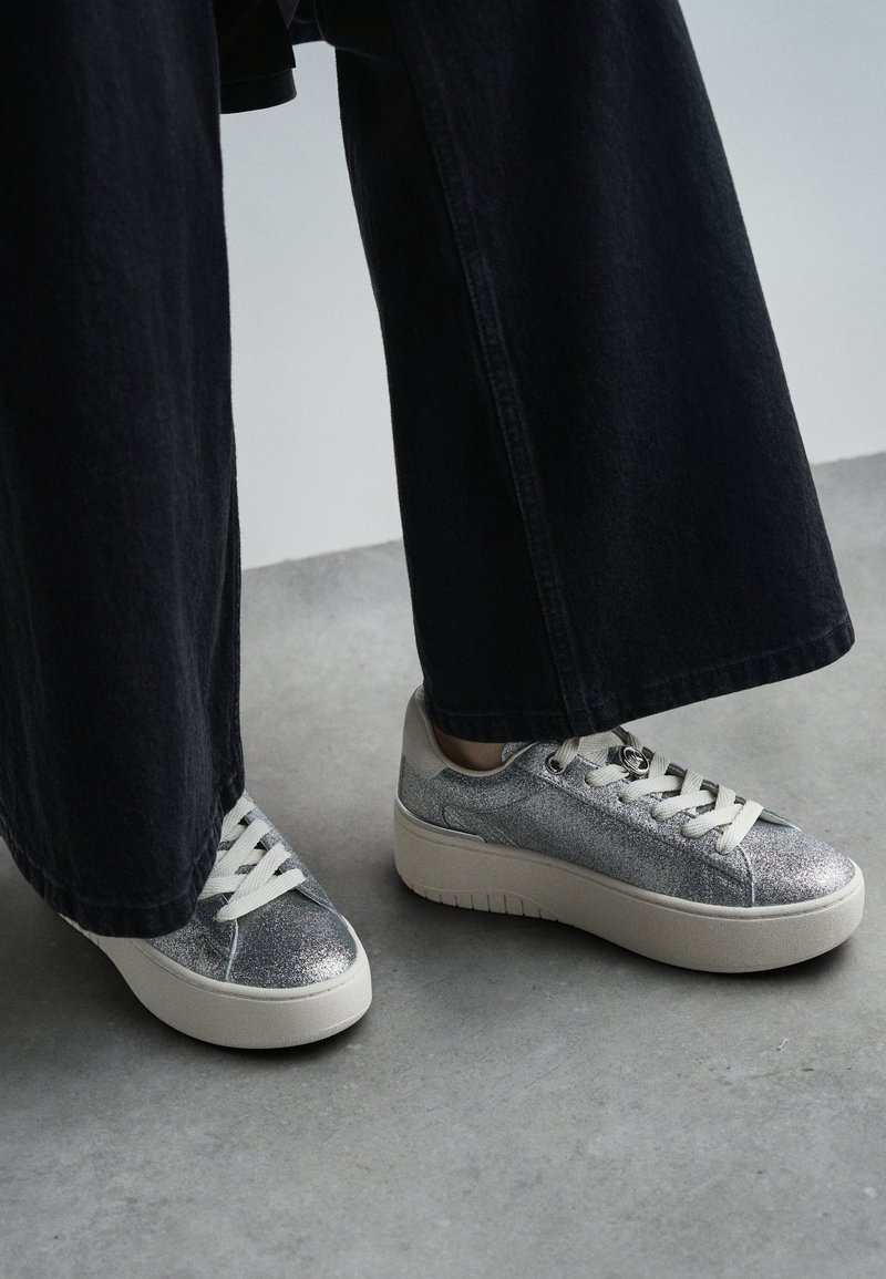 Baskets argentées pailletées avec une semelle blanche épaisse et un bout rond, équipées de lacets crème et d'œillets en métal, associées à un pantalon noir à jambes larges.