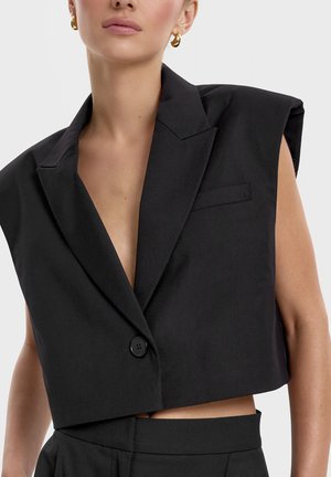 Kobieta w czarnym, bezrękawnikowym, krótko skrojonym blezerze z jednym guzikiem i złotymi kolczykami-kołami na jednolitym tle.