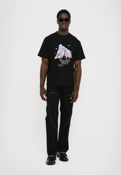 T-shirt noir avec graphique coloré de pigeon et texte. Porté avec un pantalon noir orné de clous dorés, associé à des chaussures noires brillantes.