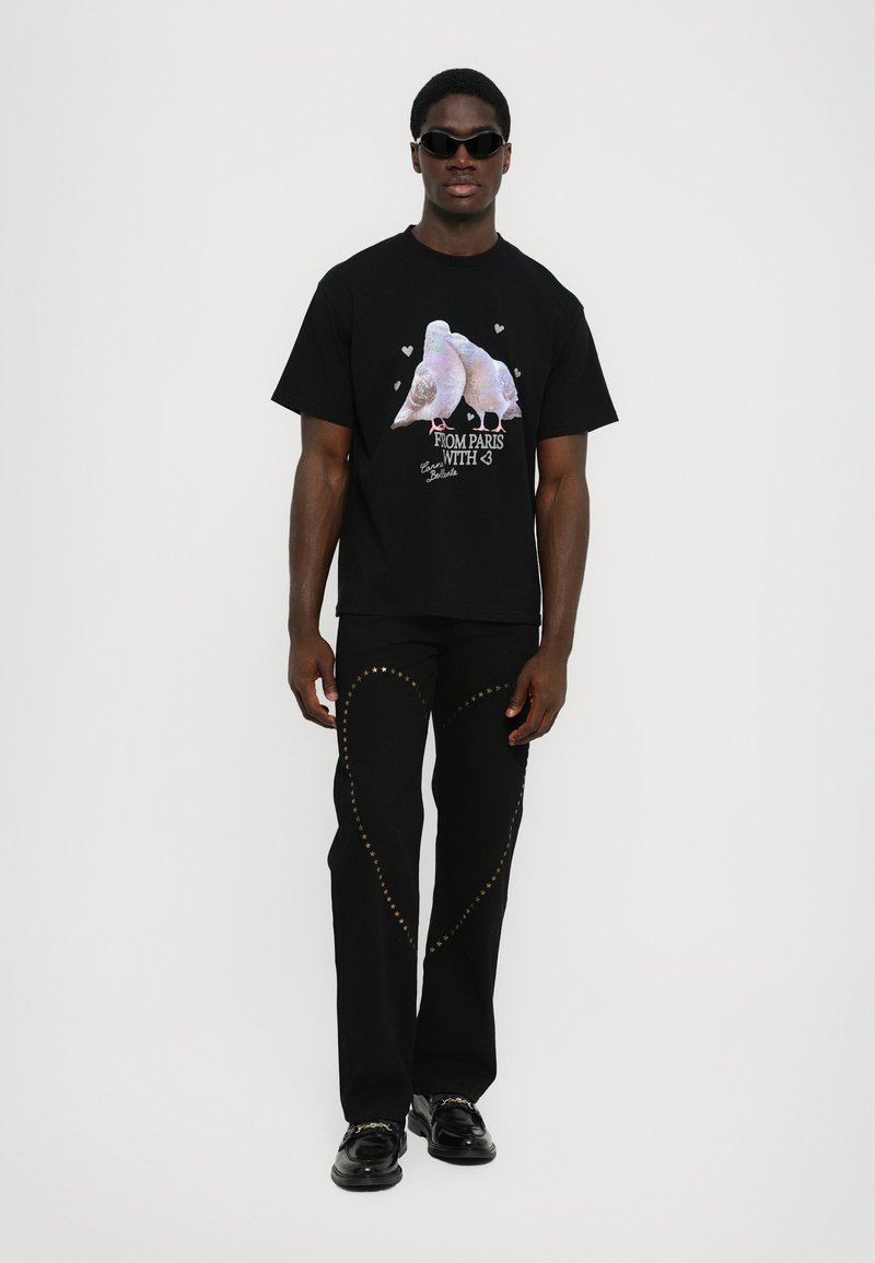 Zwarte T-shirt met kleurrijke duifgrafiek en tekst. Draagt een zwarte broek versierd met gouden studs, gecombineerd met glanzende zwarte schoenen.