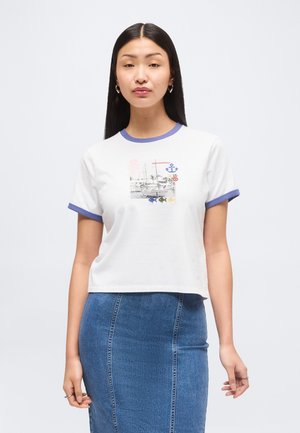 Frau mit langen dunklen Haaren, die ein weißes Ringer-T-Shirt mit Boots-Grafik sowie blaue Besätze am Kragen und an den Ärmeln trägt, kombiniert mit einem blauen Jeansrock.
