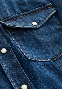 Denimskjorte detalje med en spids lomme med hvid trykknap, kontrasterende orange syninger og struktureret blå stof.