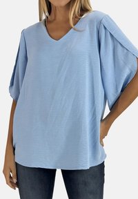 Top de manga corta azul claro en tela texturizada con escote en V y corte holgado, combinado con vaqueros oscuros. Presenta hombros caídos.