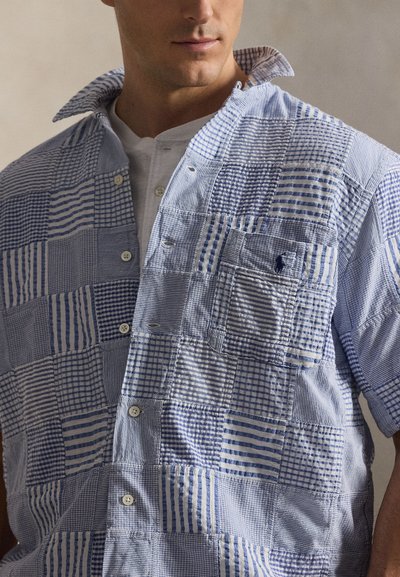 Άνδρας που φοράει μπλε και λευκό πουκάμισο κοντού μανικιού με πατσγουόρκ πάνω από λευκό πουκάμισο henley, δείχνοντας τις λεπτομέρειες γιακά και κουμπιών.