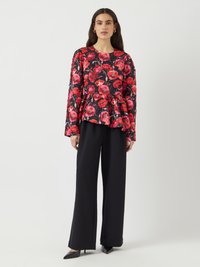 Schwarze Blumenbluse mit roten Mohnblumenmotiven, Peplum-Design, langen Ärmeln, kombiniert mit hochgeschnittenen schwarzen weiten Hosen und spitzen schwarzen Absätzen.