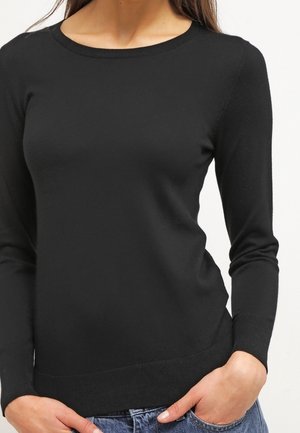 Žena s přiléhavým černým svetrem s dlouhým rukávem a kulatým výstřihem a modrými džínami, rukama v kapsách, na bílém pozadí.