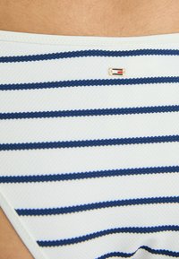 White fabric bikini bottom featuring horizontal navy blue stripes. Includes a small, branded gold accent. Textured surface with scalloped edges.