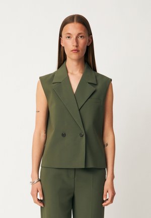 Femme aux longs cheveux bruns portant un blazer olive sans manches à double boutonnage et un pantalon assorti, debout devant un fond clair uni.