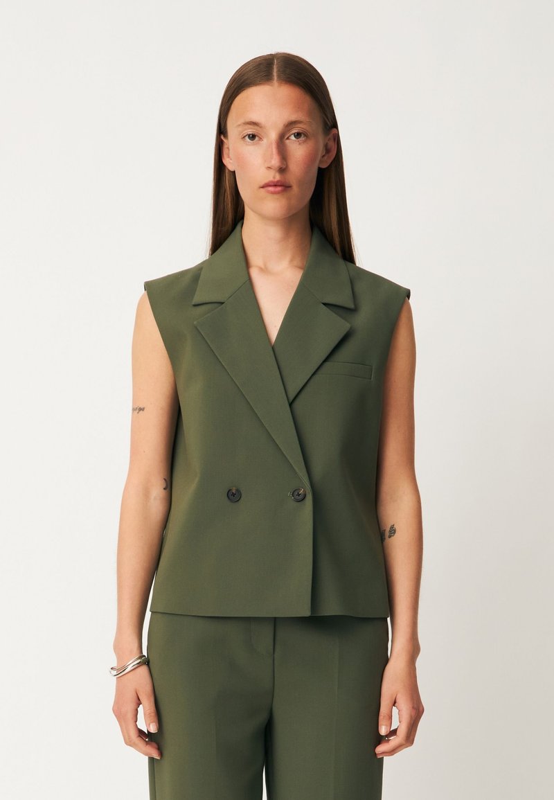 Femme aux longs cheveux bruns portant un blazer olive sans manches à double boutonnage et un pantalon assorti, debout devant un fond clair uni.