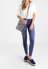 Borsa a tracolla grigia, blusa bianca con maniche, jeans skinny ricamati blu e scarpe con piattaforma navy con tomaia testurizzata.