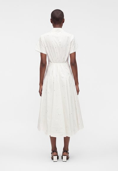 Femme aux cheveux courts portant une robe midi blanche à manches courtes avec des motifs subtils en points et des sandales noires à talons, debout de dos.