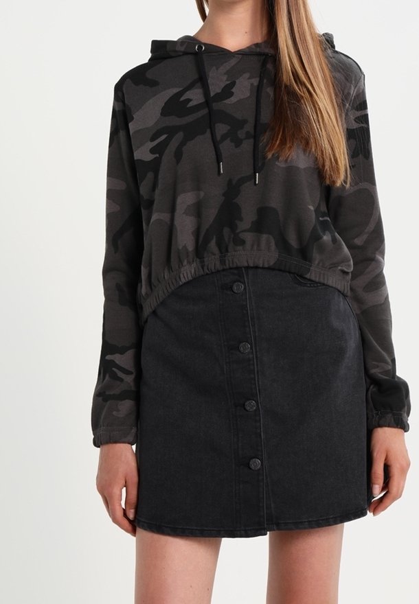 Femme portant un sweat à capuche court noir et gris camouflage associé à une jupe en jean noire taille haute avec boutons sur le devant.