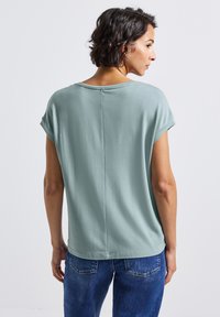 Lichtgroene blouse met korte mouwen en een ronde halslijn, gladde textuur en een middennaaddetail aan de achterkant. Gemaakt in combinatie met blauwe spijkerbroeken.