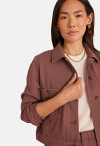 Giacca in denim marrone caratterizzata da collo con risvolto, chiusura con bottoni, taschino sul petto e dettagli sui polsini, abbinata a un top di colore chiaro.