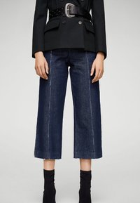 Pantalones culotte de denim azul oscuro con costuras blancas, presentando un dobladillo deshilachado. Combinados con un blazer negro ajustado y un cinturón ancho con una hebilla grande.