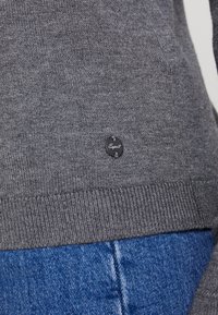 Maglione grigio lavorato a maglia con orlo a coste e un piccolo'etichetta nera con il nome del marchio "Esprit" attaccata vicino al bordo inferiore. Jeans blu visibili in basso.