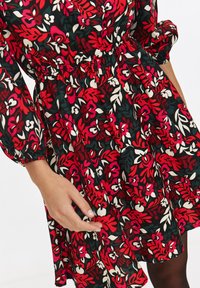 Kobieta z ręką przy biodrze, ubrana w czarną sukienkę w czerwony, biały i zielony kwiatowy wzór, z elastycznym wcięciem w talii i rękawami.