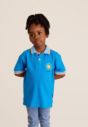 Polo bleu avec col et poignets rayés, présentant un graphique de soleil et des accents multicolores. Associé à un jean bleu clair.