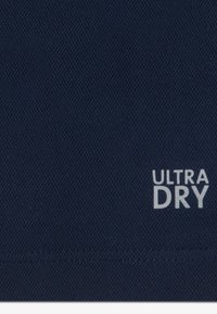 Merensininen kangas, jossa on teksturoidtu kudonta. Siinä on valkoinen "ULTRA DRY" -etiketti, joka osoittaa kosteutta siirtävät ominaisuudet.