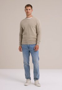 Beige mjuk tröja med rund hals, blå denimjeans och vita sneakers. Modellen står mot en neutral bakgrund. Enkel, avslappnad look.