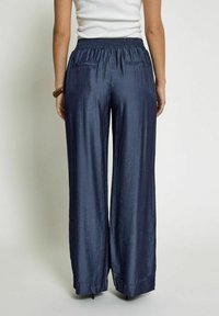Pantalon large bleu marine avec une finition texturée, une taille élastique et deux poches arrière. Le tissu drape de manière fluide, créant une silhouette décontractée.