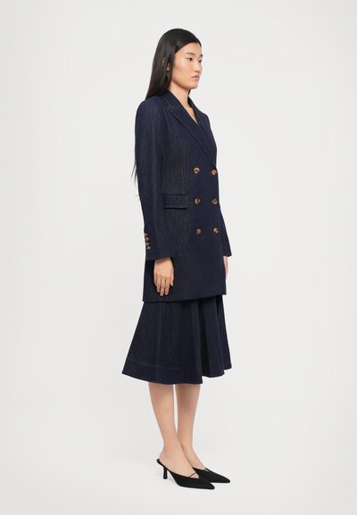 Navyblauwe dubbelgebreide blazer met gouden knopen, met een getailleerde pasvorm. Gecombineerd met een bijpassende geplisseerde rok en zwarte hakken.