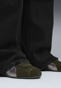 Pantalones de mezclilla oscura con dobladillos deshilachados, combinados con zapatos deportivos en verde oliva y gris metálico que presentan un acabado texturizado y un logotipo.